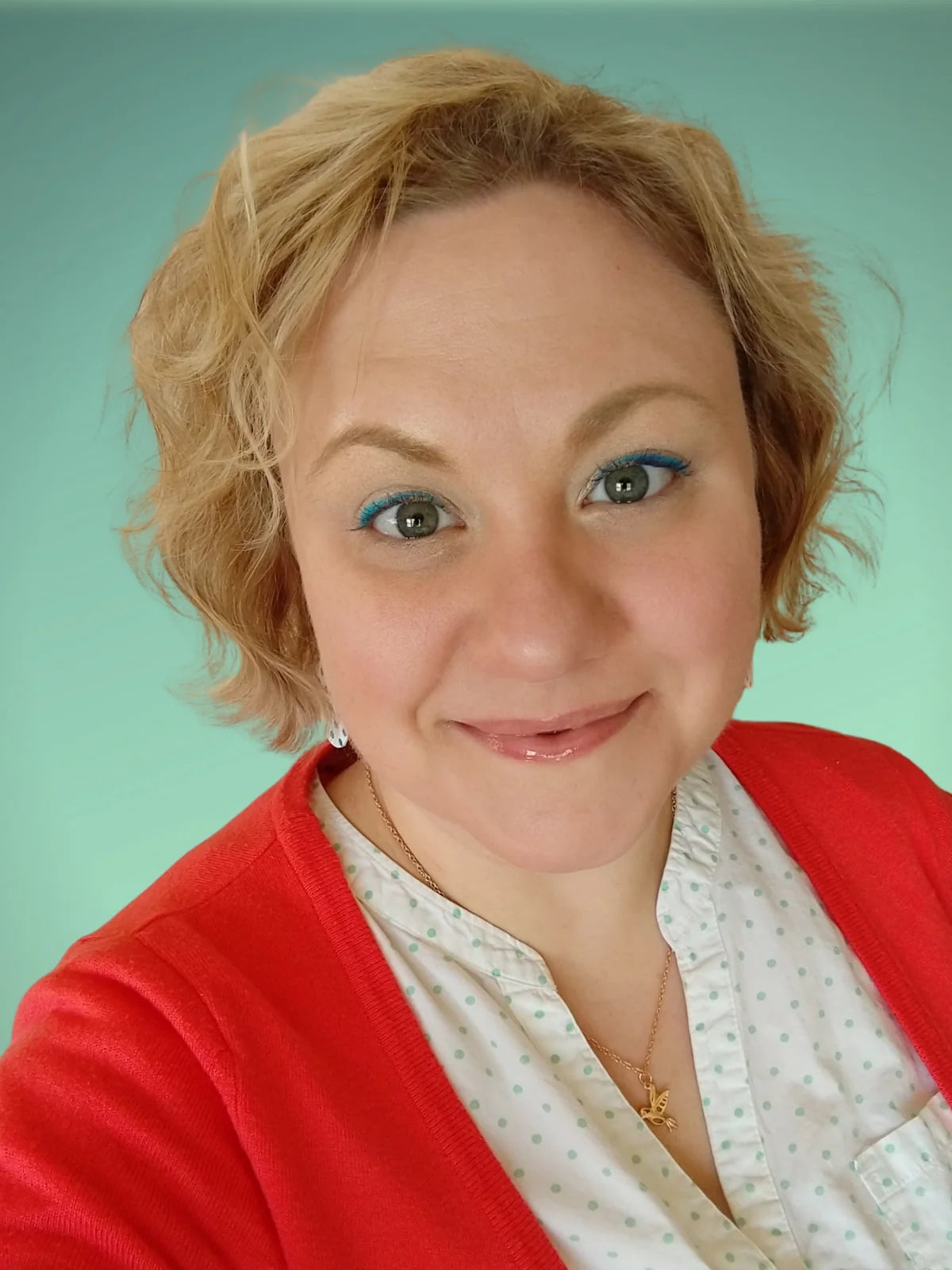 A close-up portrait of a smiling artist with short blonde hair, wearing a red cardigan over a white patterned blouse, set against a solid mint green background.