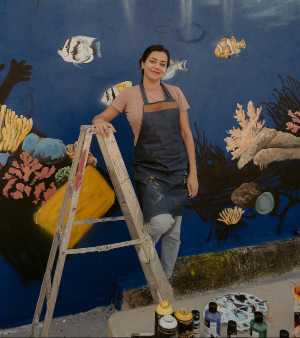 Parisa wearing a dark apron and light gray pants stands on a wooden ladder in front of a large painted mural of an underwater ocean scene with fish and coral. Paintbrushes and paint bottles are visible on a table in the foreground.